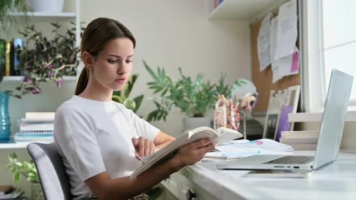 Young Woman Reading Book at Desk with Laptop