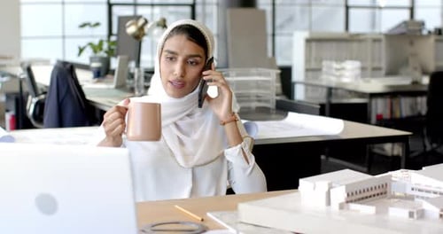 Talking on smartphone, woman working on laptop in modern office environment
