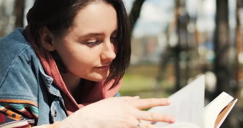 Portrait of Cheerful Young Woman Reading Book Laying on the Grass Reading Outside in Spring Sunlight