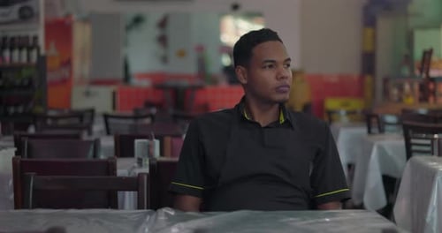 Young restaurant staff member sitting in empty dining area, looking into distance with serene and