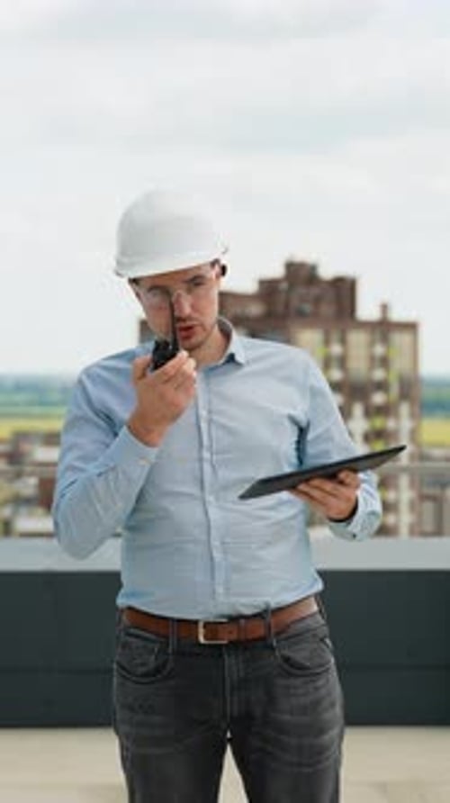 A Builder Engineer on a Construction Site Uses a Walkietalkie and a Tablet for Project Coordination