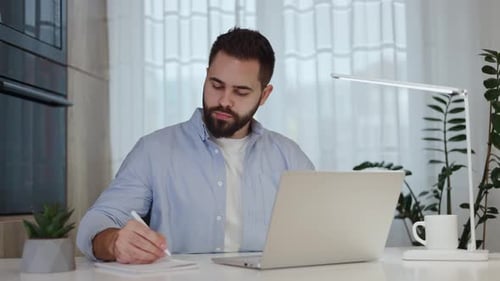 Freelancer Typing on Laptop and Writing in Notepad While Sitting at Home Office