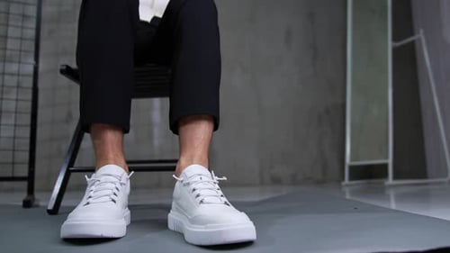 Male model in black trousers and white shirt sits on the chair. Man demonstrates white sport sneaker