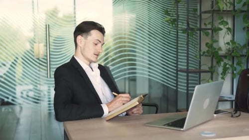 handsome, stylish young Caucasian man in business attire is sitting in a modern office, writing in a