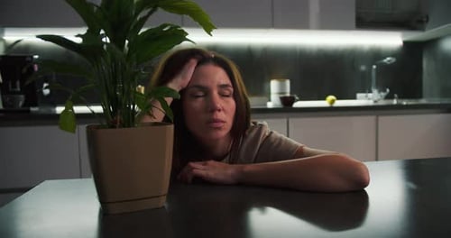 A Tired Brunette Girl in a Beige Tshirt Leans on a Black Table and Holds Her Head While Thinking