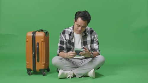 Male Traveler With Luggage Looking At The Passport And Smiling While Sitting In The Green Screen