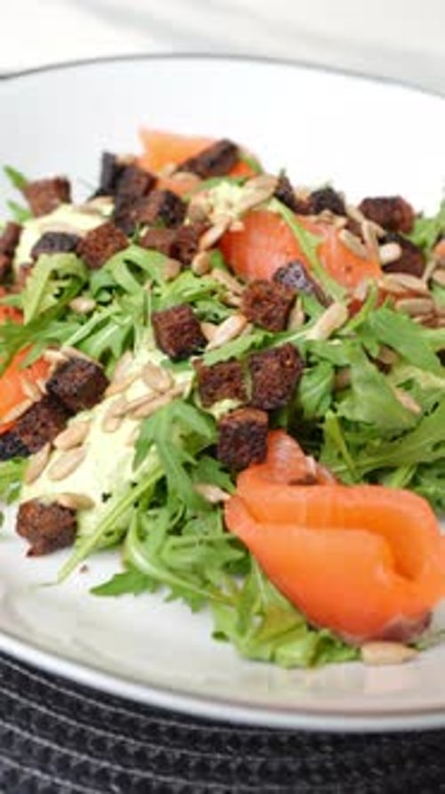 Salade de saumon rotative avec croûtons de roquette, graines de tournesol et sauce à l'avocat, servie sur un plat blanc
