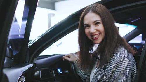 Young Woman Smiles While Sitting in a Black Car During a Sunny Indoor Event at an Automotive