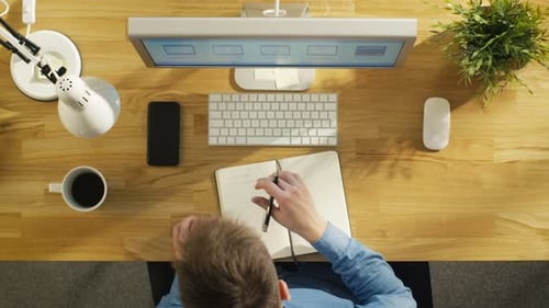 Ansicht von oben auf einen Softwareingenieur, der während der Arbeit an seinem D eine Liste mit Arbeitsthemen in sein Notizbuch schreibt