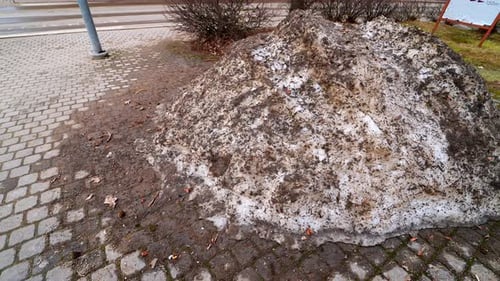 Large dirty snowbank piled beside a city sidewalk after winter, stained with sand and road grime as