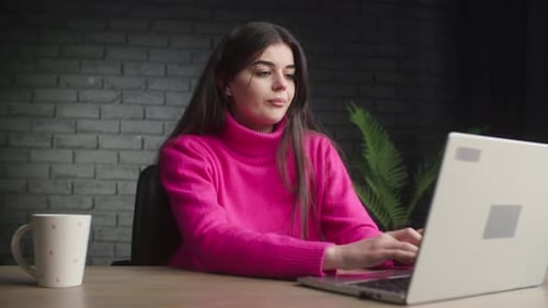 A Smiling Girl is Typing on a Laptop Young Woman in Pink Sweater Works Remotely with Computer Happy