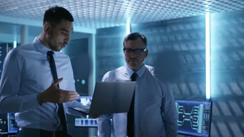 Two Security Engineers Walking In System Control Center. They Have Discussion, One of them Holds La