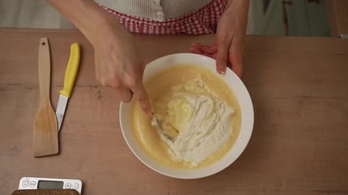 Overhead View of a Woman Baker in an Apron Mixing Flour Into Wet Batter with a Fork in a White Bowl