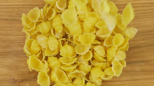 Shell-shaped pasta is poured into a glass transparent bowl against a wooden oak board. Top view.