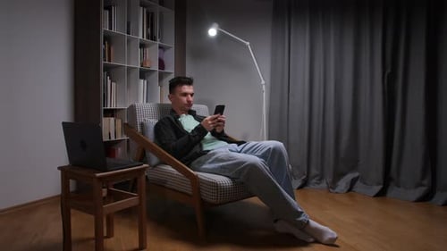 Junger Mann entspannt sich zu Hause in einem Sessel und scrollt soziale Medien und Nachrichten auf seinem Smartphone in der