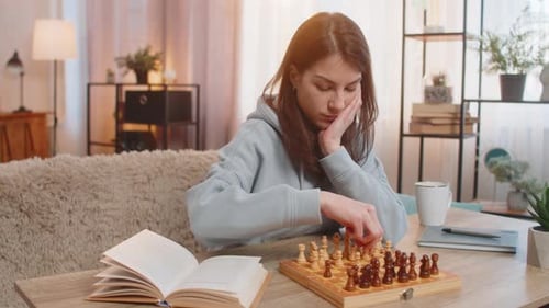 Young Woman Playing Chess Alone Arranging Pieces at Table on Sofa with Book Lying Nearby at Home