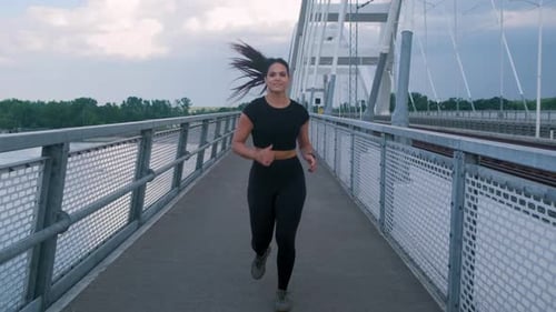 Young woman in black sport outfit running on city bridge.