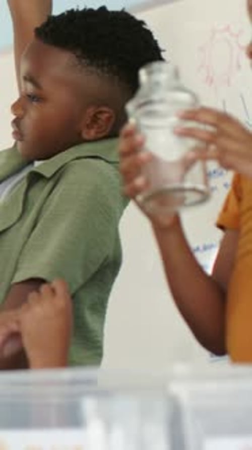 Vertical video: Reacting to teacher, orange-shirt boy handling glass jar over tub in class for demo