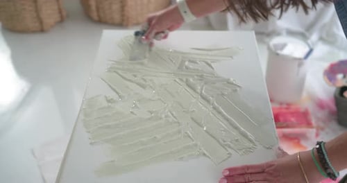 Mature woman reaching bucket spreading paste on canvas with palette knife making texture in studio