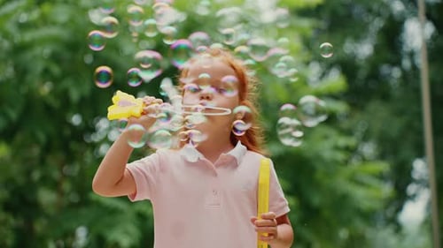 Young Girl Blows Bubbles in a Park Surrounded By Greenery on a Sunny Day
