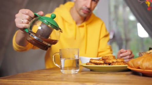 Un homme vêtu d'un sweat à capuche jaune est assis à une table dans la cuisine de sa maison et verse du thé dans une théière