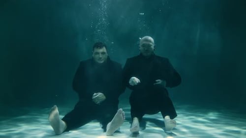 Two Men in Black Suits Sink to the Bottom of the Pool and Look at the Camera