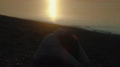 A young woman at dawn by the sea sits meditating on a yoga mat, She raises her hands up and covers t