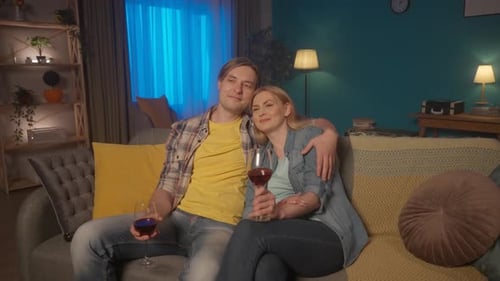 A Young Couple Sits Hugging on the Couch with Glasses of Red Wine and Watching TV A Man and a Woman