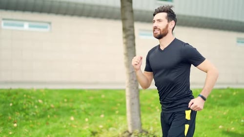 Young Caucasian bearded happy man runner jogging down the street on urban background of city park.