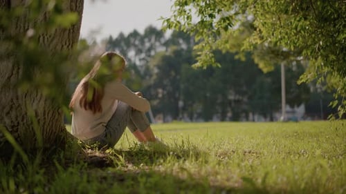 Reflective Woman Sitting on Grass in Sunlit Park with Tranquil Background