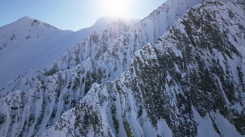 the camera flies around big snow-covered white mountain with big cliffs. the sky is blue and the sun