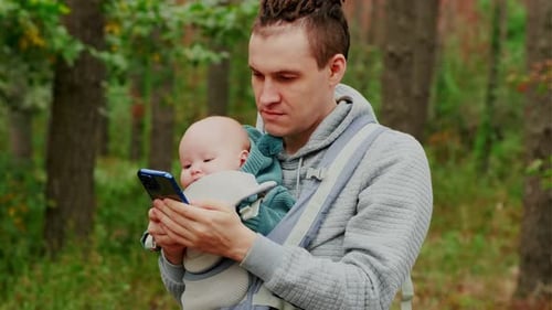 Father and Newborn Baby in a Sling in the Autumn Forest Man with Mobile Phone Happy Family