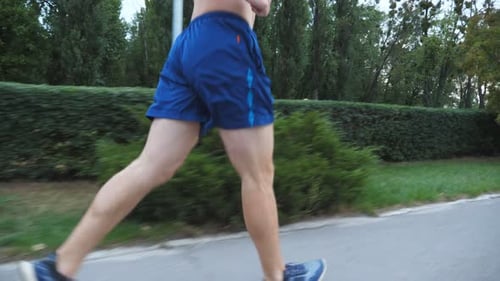 Young Muscular Sportsman with Wireless Headphones Jogging Along Sidewalk Near Forest at Summer Day