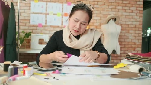 Asian female fashion designer works by choosing thread color for a dress design.