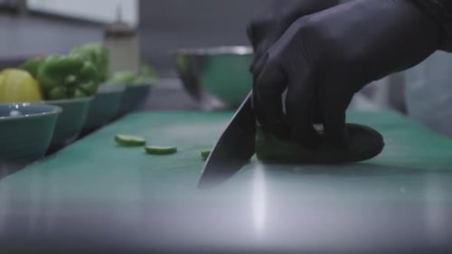 Chef cutting a cucumber using a knife with black gloves on a green cutting board. MXF