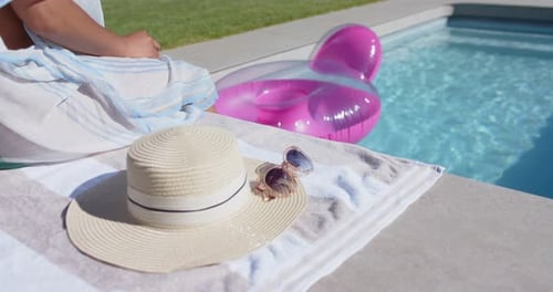 Mulher afro-americana que aquece o sol, sentada à beira da piscina, ajustando o envoltório, estendendo o pé sobre um flutuador rosa