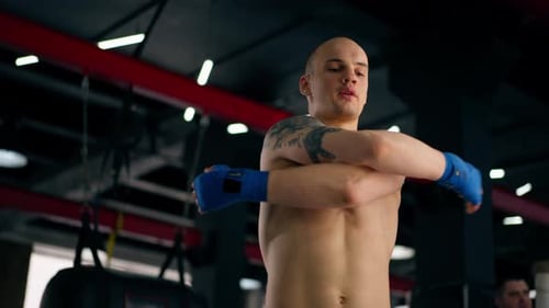 Nahaufnahme in einem Boxclub Glatzköpfiger junger Boxer mit Tattoos in blauen Bandagen, Zügen, Schlägen, Schatten