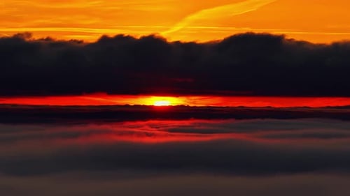 Dramatic sunrise above a cloud layer as deep orange light glows beneath a dark cloud bank, creating