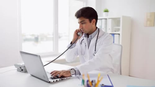 Indian doctor works on laptop and phone in hospital medical office