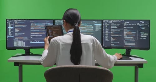 Asiatische Entwicklerin arbeitet mit Tablet, während sie Code mit mehreren Bildschirmen auf grünem Bildschirm schreibt