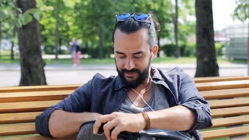 Man Sits on Park Bench Reaching into Backpack