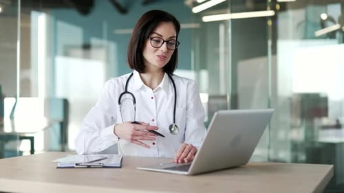 Smiling female doctor talking on video call on laptop sitting at workplace in hospital clinic