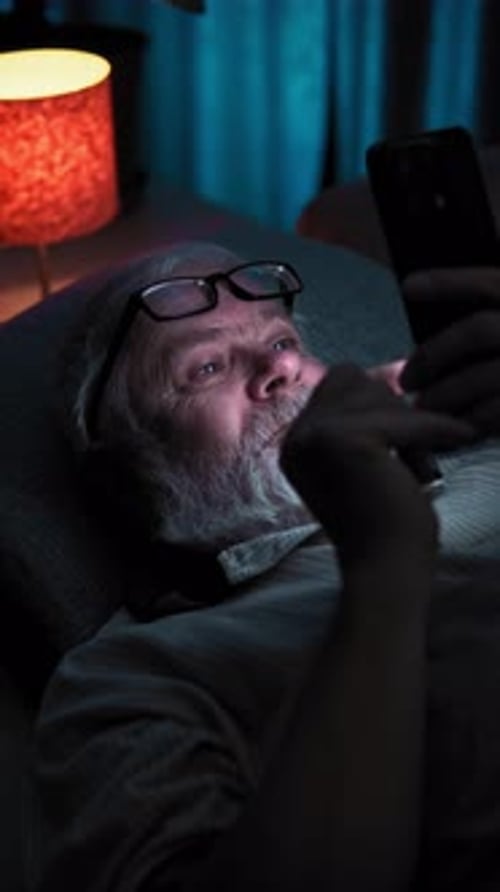 In Dark Bedroom Old Man with Glasses Lies in Bed and Surfing News on His Phone
