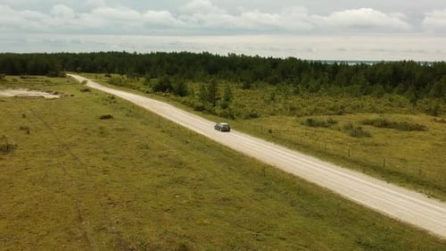Aerial shot of a grey lonely car in a dust road near the sea and a forest in a cloudy day
