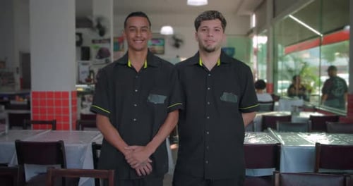 Two restaurant waiters smiling while standing inside dining area, hands folded and behind back, glas