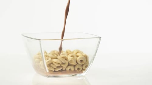 Leche con chocolate que se vierte en un recipiente transparente con cereales amarillos sobre un fondo blanco.
Desayuno