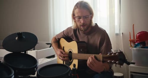 Un homme blond à lunettes avec une barbe et un t-shirt marron joue d'une guitare acoustique jaune près de lui