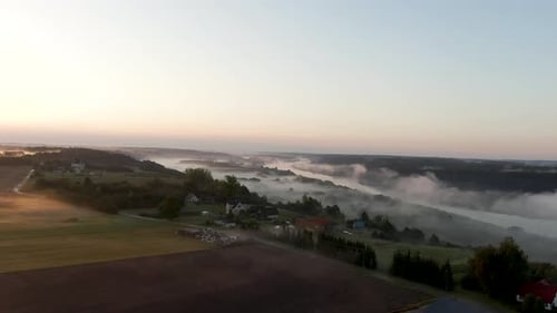 Side aerial shot of small country houses and trees while flying towards river in the foggy early mor