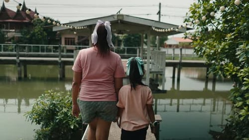 Mother and Daughter Holding Hands and Walking Together Along a Riverside Boardwalk