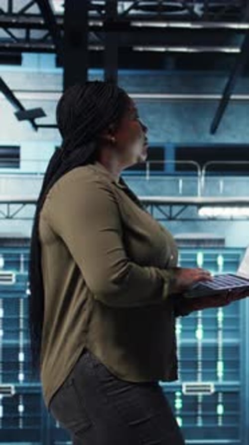 Vertical Video Engineer Walks in Server Hub Using Laptop to Upgrade Rigs